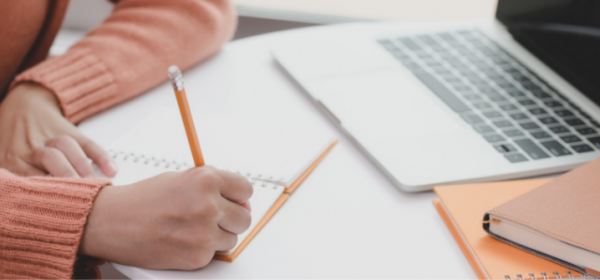 Student writing on a pad of paper with a pencil. A laptop is also on the desk.